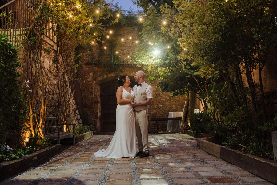 A bride and groom stand under festoon lights in a stone courtyard at dusk