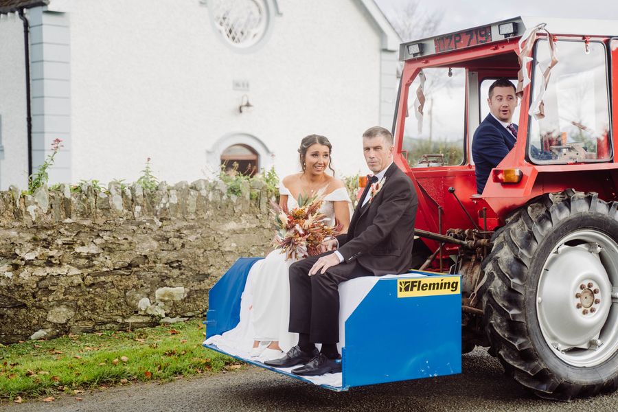 A bride and groom ride on the back of a red tractor outside the church