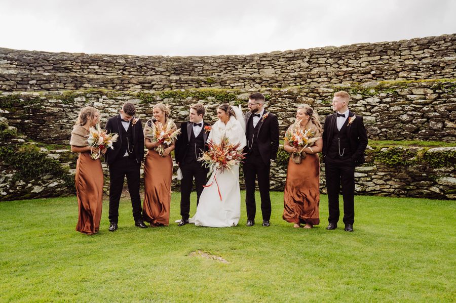 The bridal party in rust and black stand on the grass inside an old stone fort