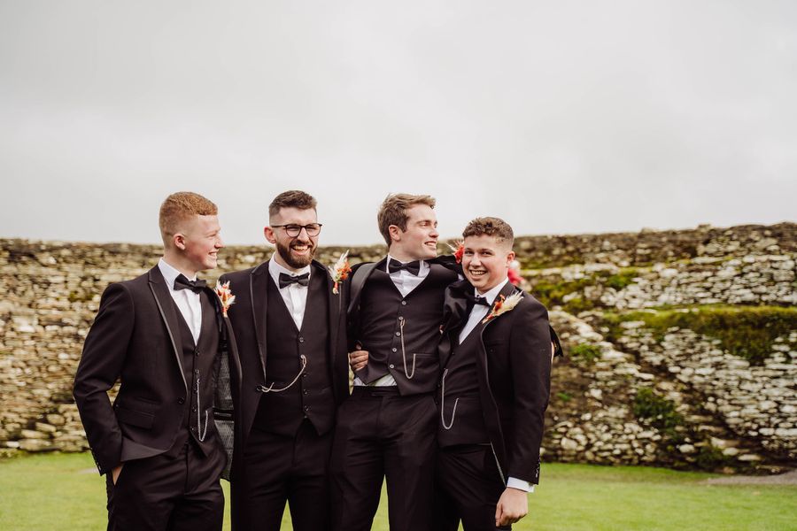 The groom and his groomsmen laugh together in front of an old stone wall
