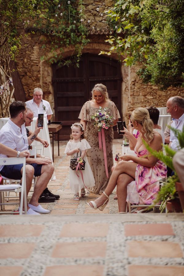 A little flower girl waits in the aisle as guests take their seats in a sunny courtyard