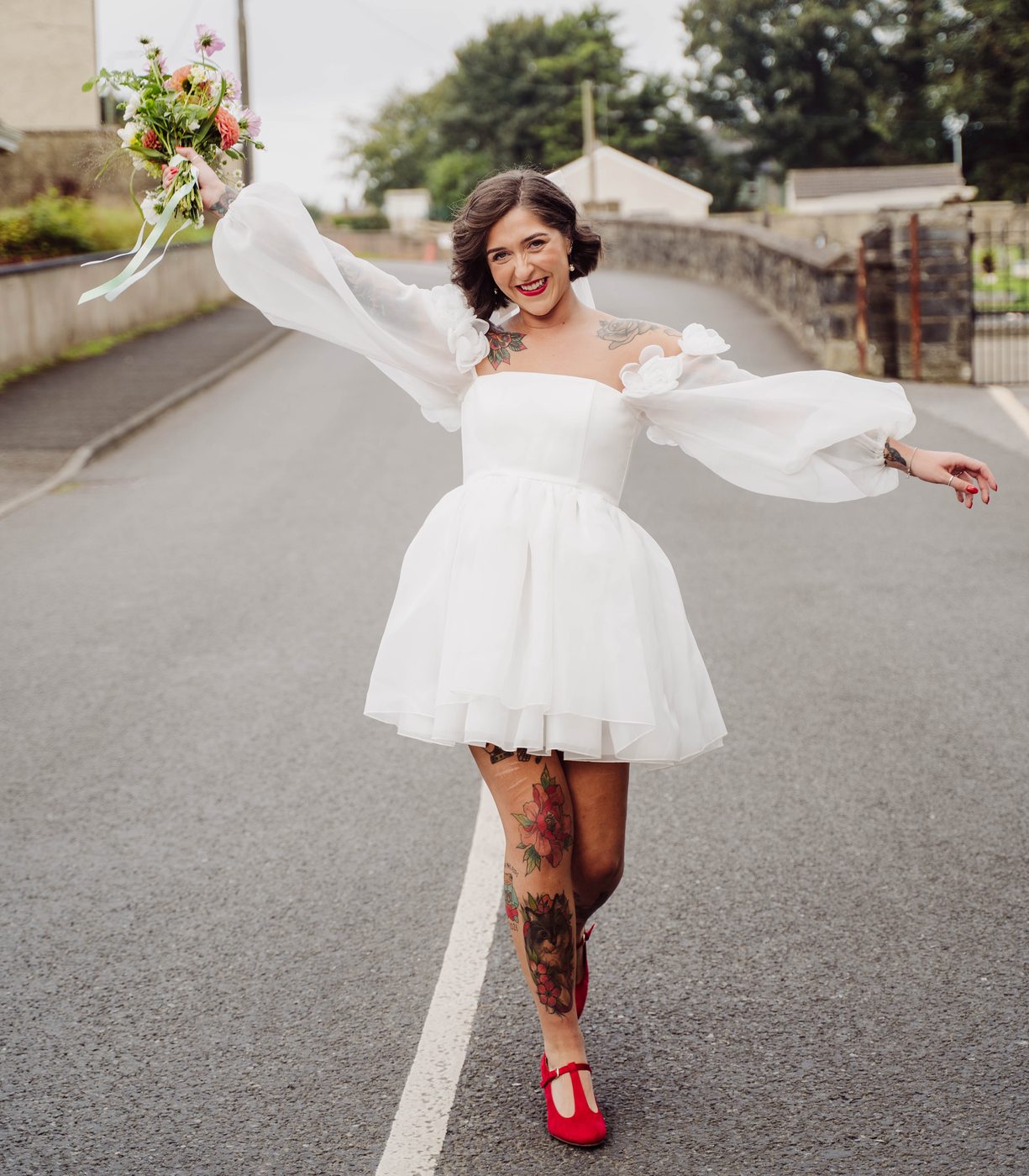 A bride in a short white dress and red shoes twirls down a country road holding her bouquet
