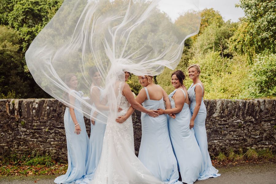 A bride's veil lifts in the wind as her bridesmaids look on