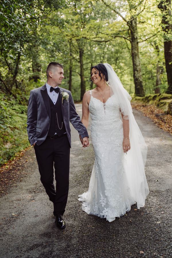 A bride and groom walk hand in hand along a woodland lane