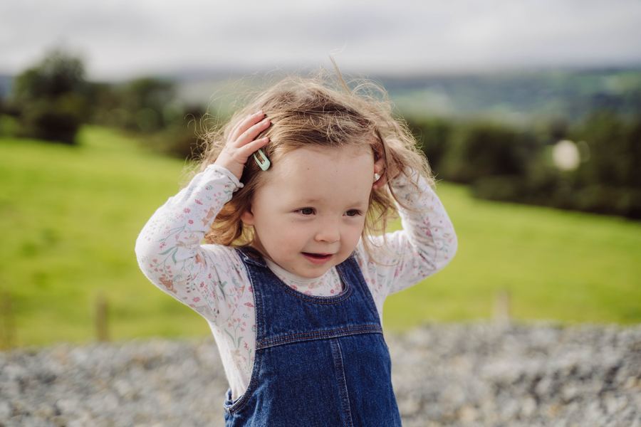 A little girl in dungarees plays with her hair in a green field