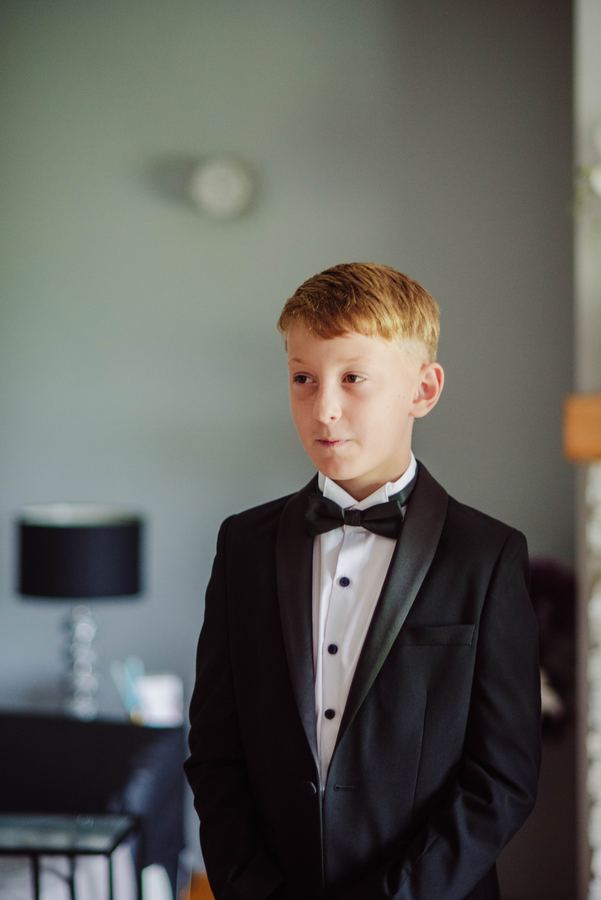A young boy in a tuxedo and bow tie waits by the window